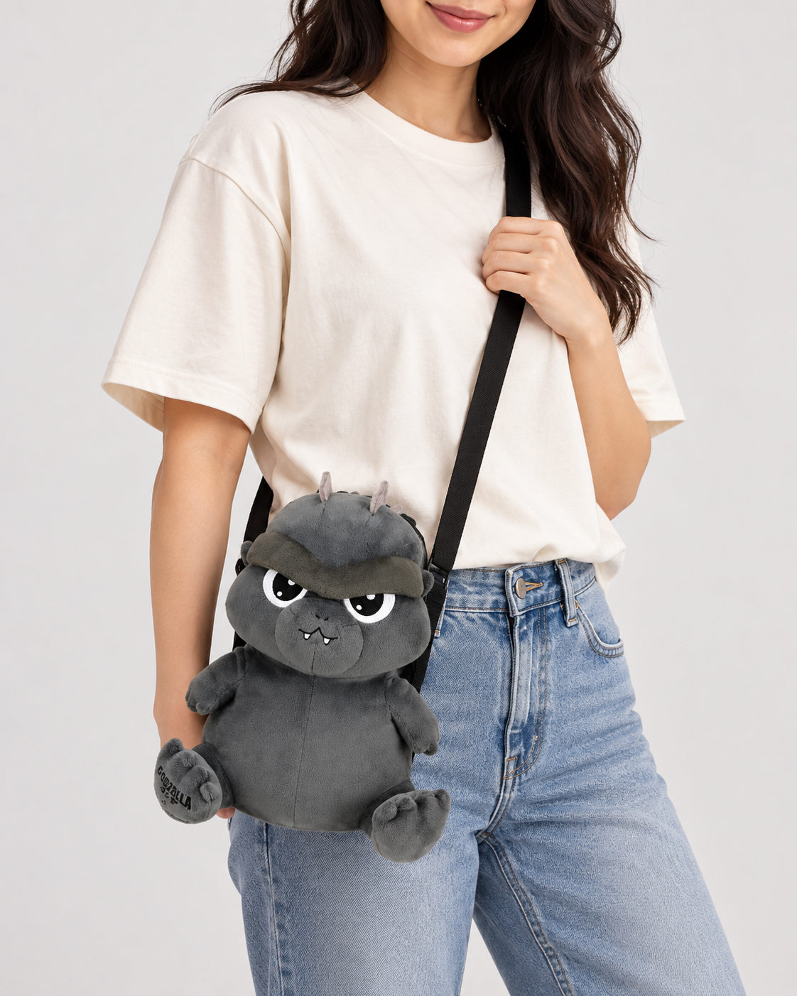 A woman wearing a cream t-shirt and blue jeans carries a dark gray plush Godzilla crossbody bag with a black strap against a light gray studio background.