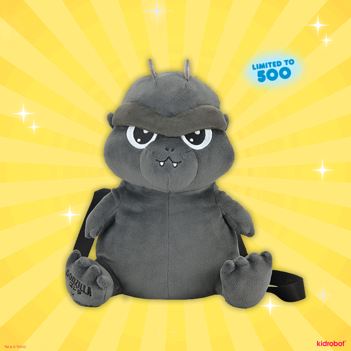 A gray plush Godzilla backpack with a black strap, shown against a bright yellow sunburst background with sparkles and a blue 'LIMITED TO 500' badge. Kidrobot branding is visible in the bottom right corner.