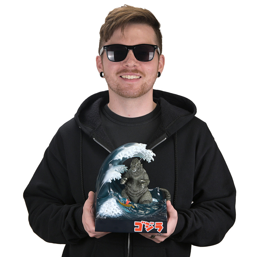 A man wearing a black hoodie and sunglasses holds up a Godzilla collectible figure inside a wave-themed display dome. The figure depicts Godzilla rising from the ocean with a small Dunny in a Boat, and the word 'ゴジラ' is printed in red at the bottom of the display.